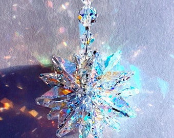 Peut inclure: Un attrape-soleil en cristal avec un motif en étoile éclate suspendu à une chaîne de perles de cristal claires. L'attrape-soleil projette des arcs-en-ciel sur une surface blanche.