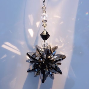 May include: A black crystal suncatcher with a black crystal pendant hanging from a black string. The suncatcher is made of many small crystals that are arranged in a star shape. The pendant is a single, large crystal that is shaped like a diamond.