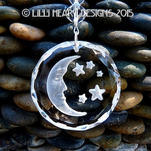 May include: A clear crystal suncatcher with a crescent moon and stars etched into it. The suncatcher is hanging from a silver chain and is surrounded by smooth, gray stones.