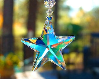 May include: A crystal star suncatcher with a rainbow iridescent finish hangs from a string of clear and red crystal beads. The star is suspended in front of a blurred background of green trees.