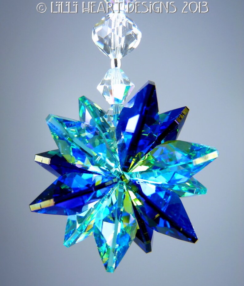 May include: A faceted, star-shaped crystal ornament in shades of blue and green, suspended by a clear beaded strand. The ornament's multiple facets reflect light, creating a sparkling effect. The text "&copy; LILLI HEART DESIGNS 2013" is visible at the top.