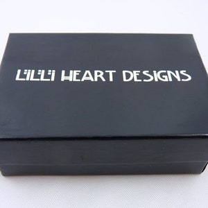Puede incluir: Una caja negra con el texto "Lilli Heart Designs" impreso en blanco.
