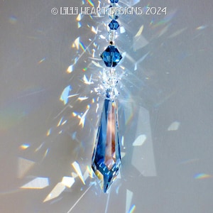 May include: A teardrop-shaped crystal prism hangs from a string of smaller crystals. The prism is a deep blue color and casts rainbows on the white background.