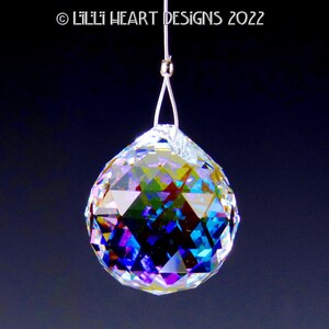 Puede incluir: Una bola de cristal transparente con colores arco&iacute;ris iridiscentes que cuelga de una cuerda plateada. La bola es facetada y refleja la luz.
