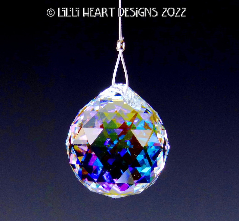 Puede incluir: Una bola de cristal transparente con colores arco&iacute;ris iridiscentes que cuelga de una cuerda plateada. La bola es facetada y refleja la luz.
