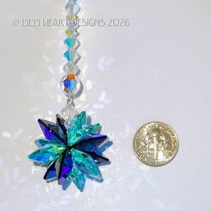May include: A crystal suncatcher with a starburst design, featuring shades of teal, blue, and purple. The suncatcher is suspended by a string of clear and iridescent beads. A US dime is next to the suncatcher for size reference. The text "&copy; Lilii Heart Designs 2026" is in the upper left corner.