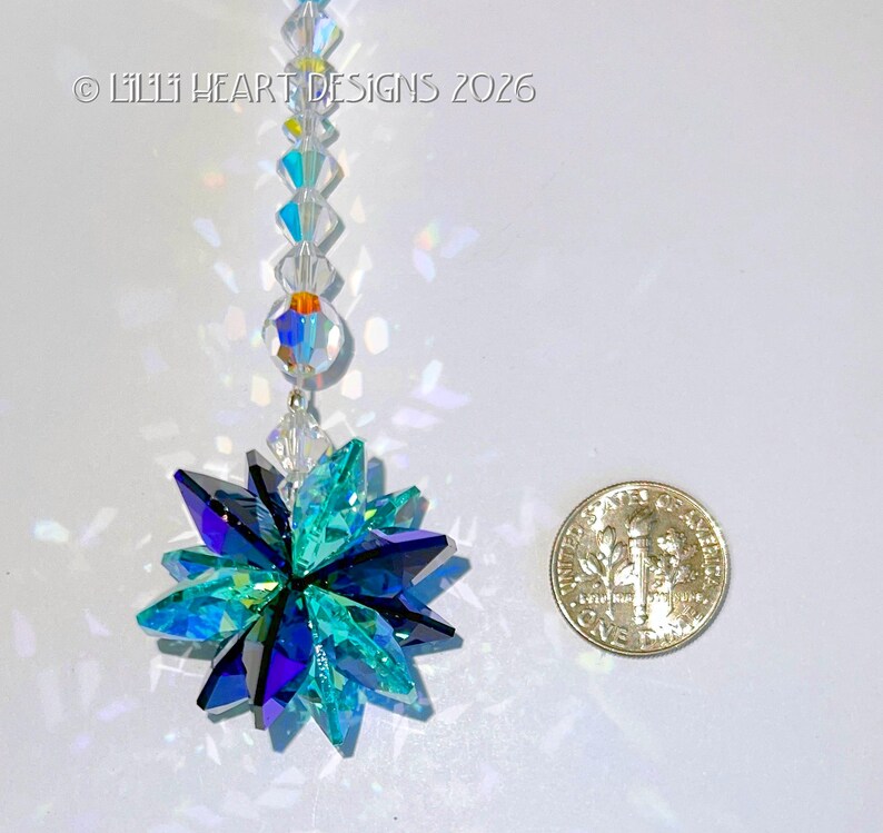 May include: A crystal suncatcher with a starburst design, featuring shades of teal, blue, and purple. The suncatcher is suspended by a string of clear and iridescent beads. A US dime is next to the suncatcher for size reference. The text "&copy; Lilii Heart Designs 2026" is in the upper left corner.