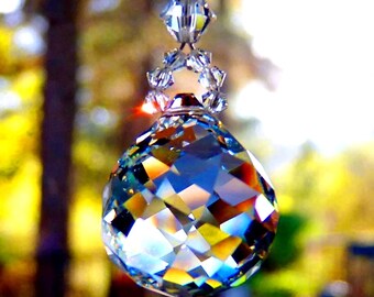 Op de afbeelding: Een kristallen zonenvanger met een grote, gefacetteerde, iriserende bal die aan een zilveren ketting hangt. De bal reflecteert regenboogkleuren. De zonenvanger hangt voor een wazige achtergrond van groen en geel.