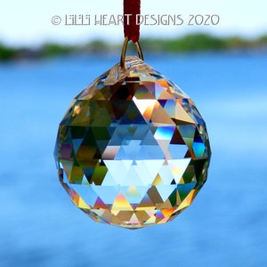 May include: A clear crystal ball with a faceted design hangs from a gold wire and red string. The ball is reflecting light, creating a rainbow effect.