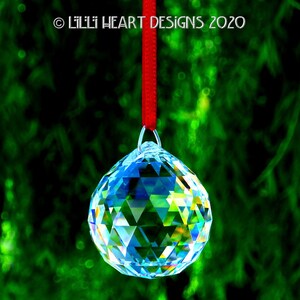 May include: A clear crystal ball hanging from a red ribbon against a green background. The crystal ball is faceted and reflects light.