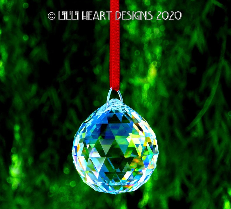 May include: A clear crystal ball hanging from a red ribbon against a green background. The crystal ball is faceted and reflects light.