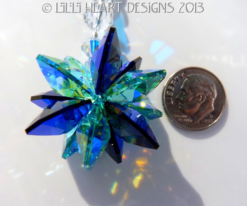 May include: A faceted crystal starburst ornament in shades of blue and green, with a silver dime for size comparison. The ornament is suspended from a clear beaded strand. The image is well-lit, showcasing the crystal's reflective qualities.