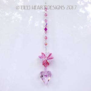 May include: Pink crystal suncatcher with a heart-shaped crystal at the bottom and a flower-shaped crystal cluster in the middle. The suncatcher is hanging from a white ceiling.