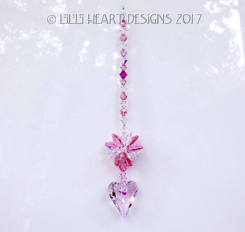 May include: Pink crystal suncatcher with a heart-shaped crystal at the bottom and a flower-shaped crystal cluster in the middle. The suncatcher is hanging from a white ceiling.