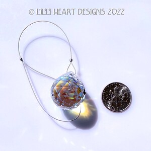 Puede incluir: Una bola de cristal transparente con facetas iridiscentes cuelga de un lazo de alambre plateado. La bola de cristal es aproximadamente del tama&ntilde;o de una moneda de diez c&eacute;ntimos de euro.