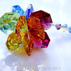 Puede incluir: Un amuleto de flor de cristal colorido con un arco&iacute;ris de colores, incluyendo rojo, naranja, amarillo, verde, azul y morado. El amuleto est&aacute; unido a una cadena plateada con cristales m&aacute;s peque&ntilde;os.