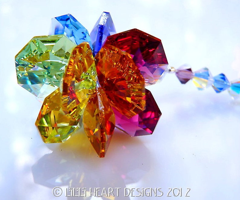 Puede incluir: Un amuleto de flor de cristal colorido con un arco&iacute;ris de colores, incluyendo rojo, naranja, amarillo, verde, azul y morado. El amuleto est&aacute; unido a una cadena plateada con cristales m&aacute;s peque&ntilde;os.