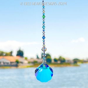 May include: A hanging crystal suncatcher with a faceted, teardrop-shaped pendant in shades of blue. The suncatcher is strung with a series of colorful beads, including clear, green, and blue. The text "© Lilli Heart Designs 2024" is visible at the top.
