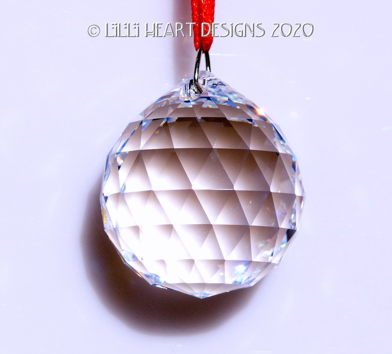 May include: A clear crystal ball with a faceted surface hangs from a red string.  The ball is approximately 2 inches in diameter.  &copy; Lilli Heart Designs 2020