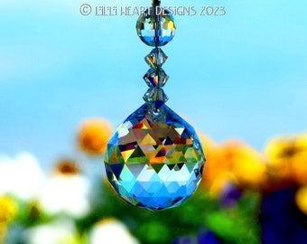 Puede incluir: Un atrapasol de cristal con una gran bola azul facetada que cuelga de una cadena de cuentas de cristal más pequeñas. El atrapasol está colgado frente a un fondo borroso de flores amarillas y blancas.