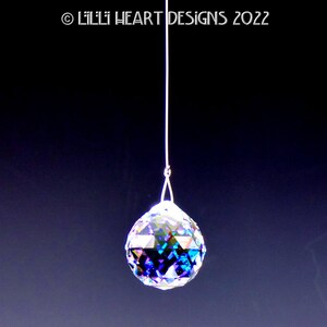 Puede incluir: Una bola de cristal colgando de una cuerda blanca. La bola es iridiscente y tiene un dise&ntilde;o facetado. Est&aacute; colgando sobre un fondo azul oscuro.