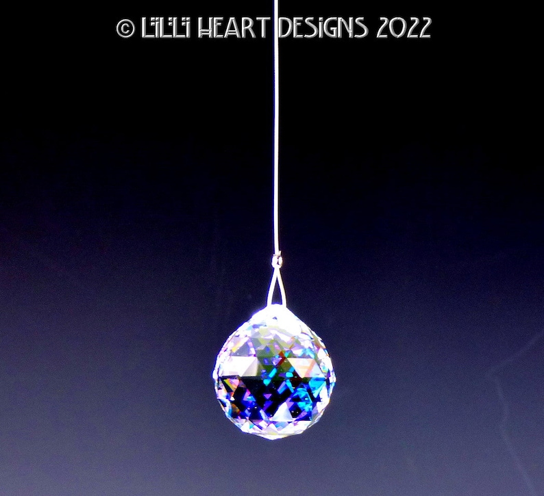 Puede incluir: Una bola de cristal colgando de una cuerda blanca. La bola es iridiscente y tiene un dise&ntilde;o facetado. Est&aacute; colgando sobre un fondo azul oscuro.