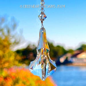 以下が含まれることがあります： 透明なクリスタルのサンキャッチャー。多面的なデザインで、透明なビーズのチェーンから吊り下げられています。サンキャッチャーは幾何学的なベースを持つ涙の形をしています。背景には、水面と葉のぼやけた景色が写っています。
