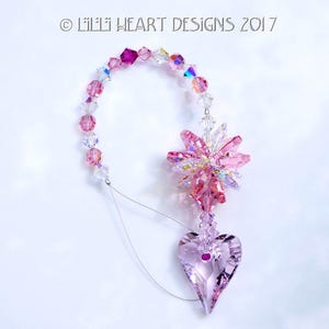May include: Pink crystal suncatcher with a heart-shaped crystal pendant. The suncatcher is made of pink and clear crystals and is strung on a silver wire. The heart-shaped pendant is also made of pink crystal and has a small, clear crystal in the center.