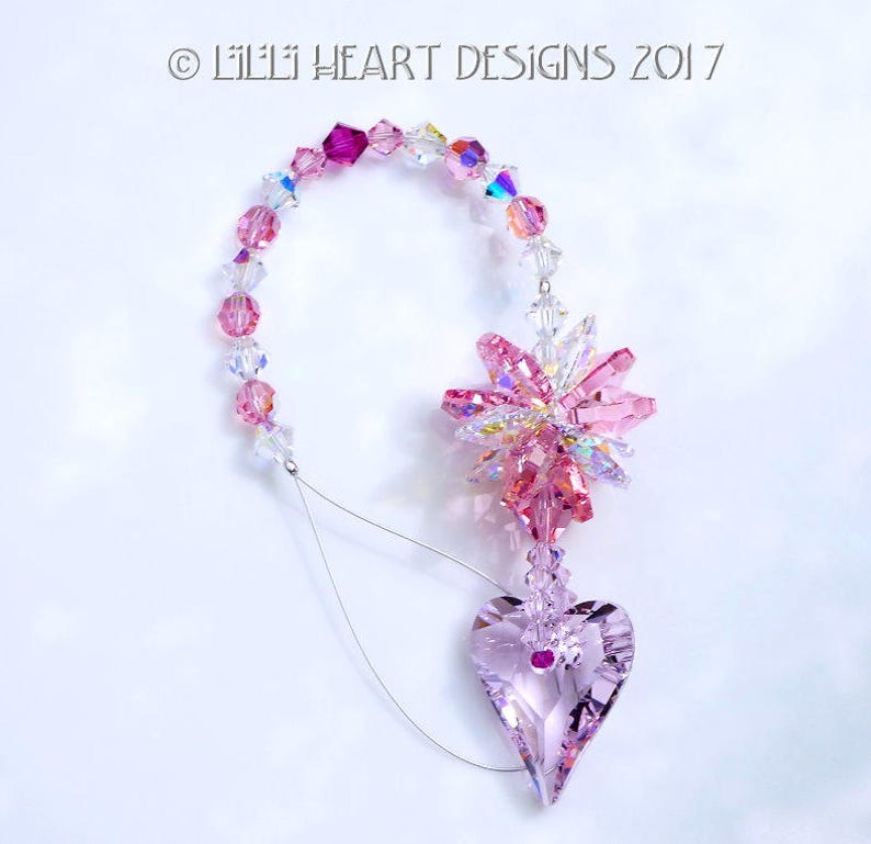 May include: Pink crystal suncatcher with a heart-shaped crystal pendant. The suncatcher is made of pink and clear crystals and is strung on a silver wire. The heart-shaped pendant is also made of pink crystal and has a small, clear crystal in the center.