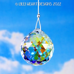 Puede incluir: Una bola de cristal transparente colgando de una cuerda blanca sobre un fondo de cielo azul. La bola de cristal es facetada y refleja colores arco&iacute;ris.