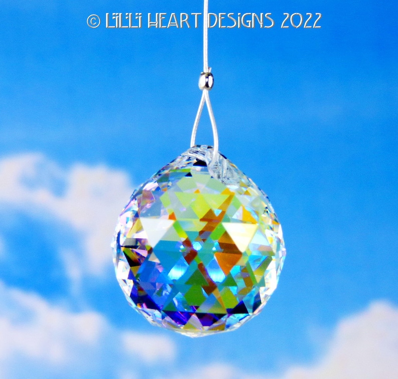 Puede incluir: Una bola de cristal transparente colgando de una cuerda blanca sobre un fondo de cielo azul. La bola de cristal es facetada y refleja colores arco&iacute;ris.