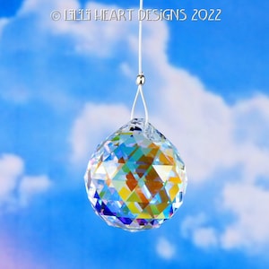 Puede incluir: Una bola de cristal transparente colgando de una cuerda blanca contra un cielo azul con nubes blancas. La bola de cristal es facetada y refleja colores arco&iacute;ris.