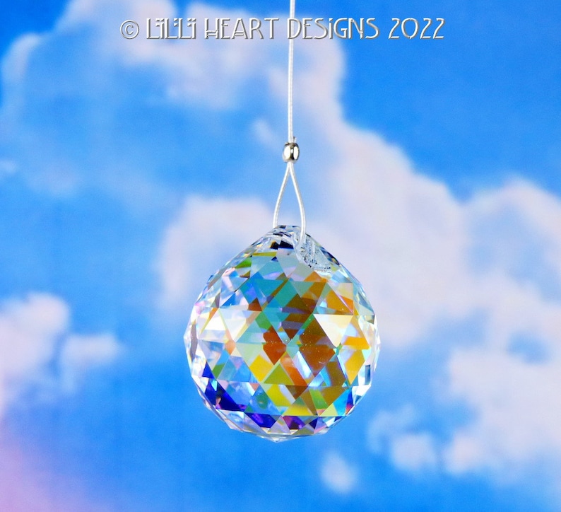 Puede incluir: Una bola de cristal transparente colgando de una cuerda blanca contra un cielo azul con nubes blancas. La bola de cristal es facetada y refleja colores arco&iacute;ris.
