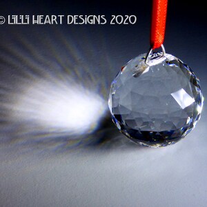 May include: A clear crystal ball hanging from a red ribbon. The ball casts a rainbow-like reflection on the surface below.