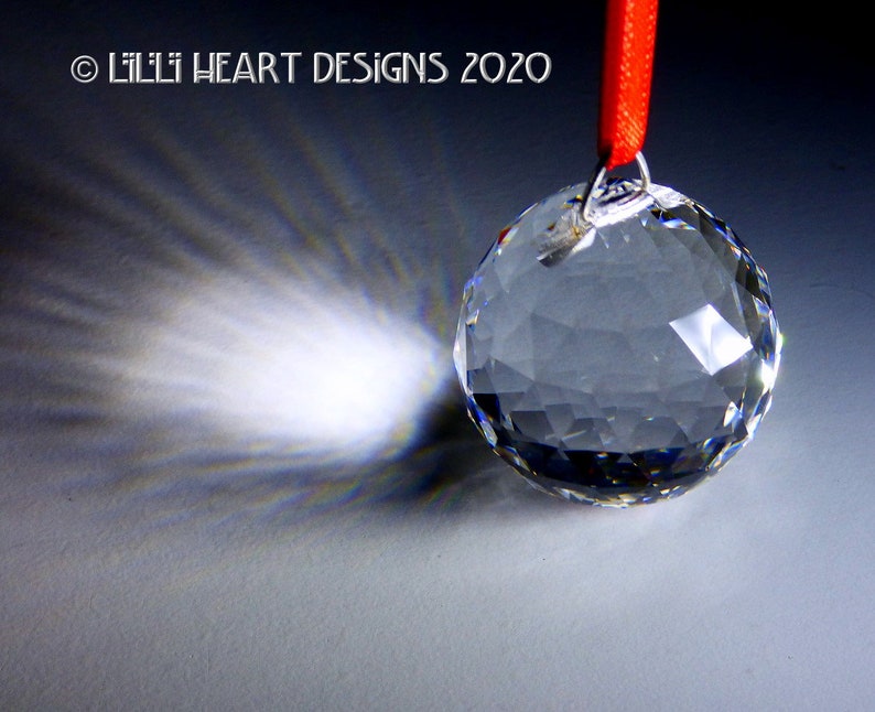 May include: A clear crystal ball hanging from a red ribbon. The ball casts a rainbow-like reflection on the surface below.