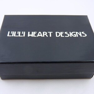 Puede incluir: Una caja negra con el texto "Lilli Heart Designs" impreso en blanco.