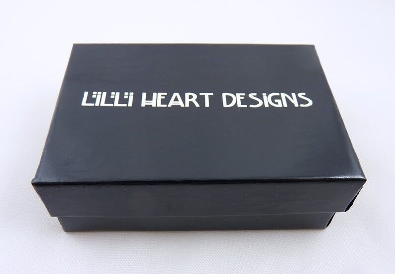 Puede incluir: Una caja negra con el texto "Lilli Heart Designs" impreso en blanco.