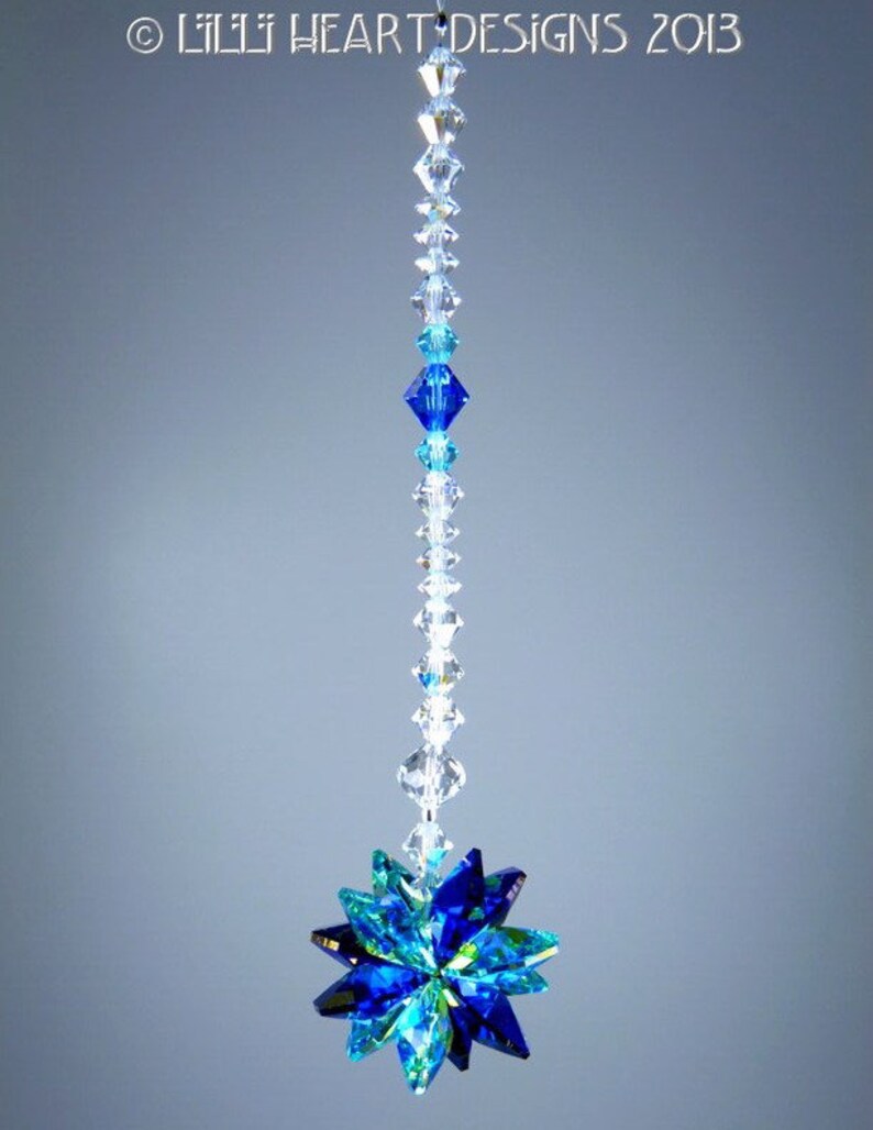 May include: A hanging crystal ornament with a starburst design. The ornament features a string of clear and blue crystals leading to a multi-faceted starburst in shades of blue and green. The text "&copy; Lilli Heart Designs 2013" is visible.