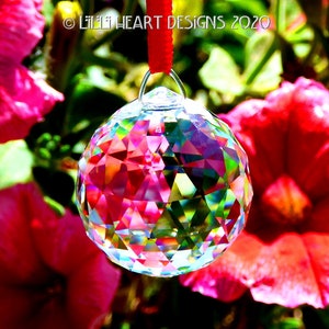 May include: A clear crystal ball with rainbow facets hangs from a red ribbon against a background of pink flowers.