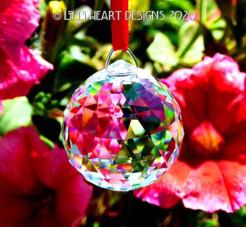 May include: A clear crystal ball with rainbow facets hangs from a red ribbon against a background of pink flowers.