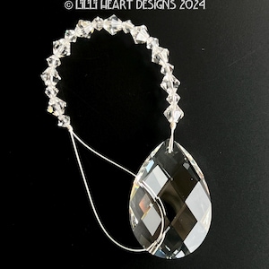 Puede incluir: Un colgante de cristal transparente en forma de lágrima con una cadena de cuentas de cristal. La cadena está unida al colgante con un lazo de alambre plateado.