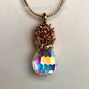 May include: A gold-colored pineapple pendant necklace with a faceted, iridescent crystal base. The pendant is suspended from a delicate gold chain. The text "© Lili Heart Designs 2026" is visible.