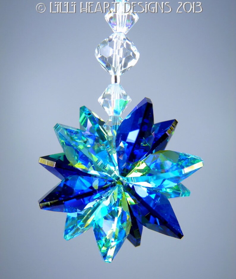 May include: A hanging crystal ornament with a starburst design. The ornament features faceted blue and green crystals arranged in a star shape, with a clear crystal strand above. The text "&copy; LILLI HEART DESIGNS 2013" is at the top.