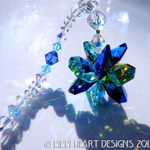 May include: A decorative crystal suncatcher featuring a faceted flower design in shades of blue and green. The flower is suspended from a strand of clear, iridescent beads. The image includes the text "&copy; LILII HEART DESIGNS 2013".