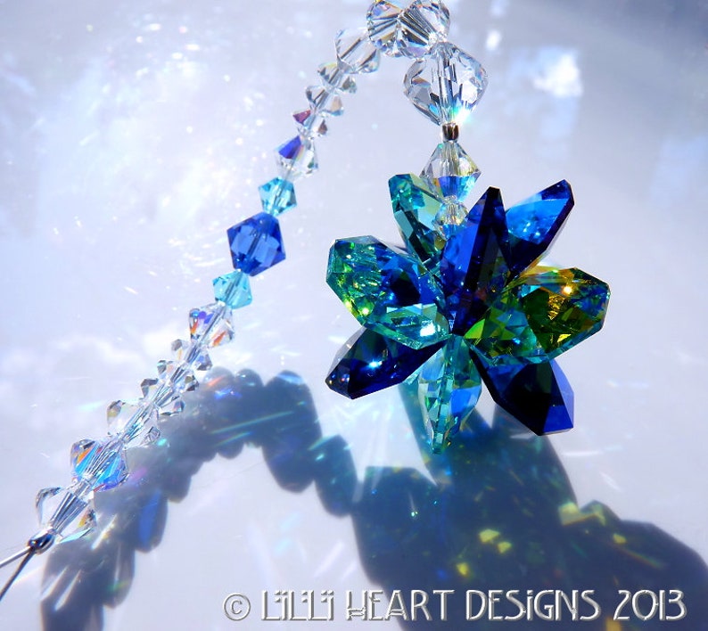 May include: A decorative crystal suncatcher featuring a faceted flower design in shades of blue and green. The flower is suspended from a strand of clear, iridescent beads. The image includes the text "&copy; LILII HEART DESIGNS 2013".