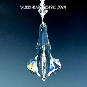 May include: A clear, faceted crystal pendant with a decorative shape, suspended by a thin silver-colored wire. The crystal reflects a rainbow of colors. The text "© Lilli Heart Designs 2024" is visible at the top.