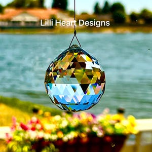 May include: A faceted crystal ball, suspended by a black string, reflects light with a rainbow of colors. The text "Lilli Heart Designs" is visible in the background. The crystal ball is a decorative item.