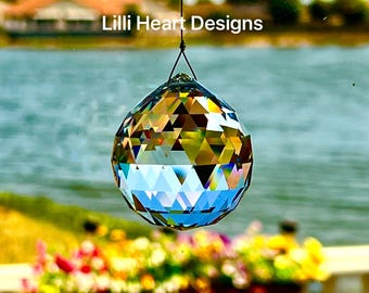 Puede incluir: Una bola de cristal facetada, suspendida por un hilo negro, refleja la luz con un arcoíris de colores. El texto "Lilli Heart Designs" es visible. Es un artículo decorativo.