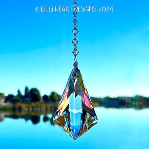 May include: A clear, faceted crystal prism pendant hangs from a chain against a blue sky and water background. The prism refracts light, displaying a spectrum of colors. Text at the top reads "© LILI HEART DESIGNS 2024".