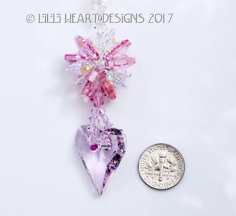 May include: A pink and clear crystal suncatcher with a heart-shaped crystal pendant. The suncatcher is hanging from a silver chain. The image includes the text "Lilli Heart Designs 2017".